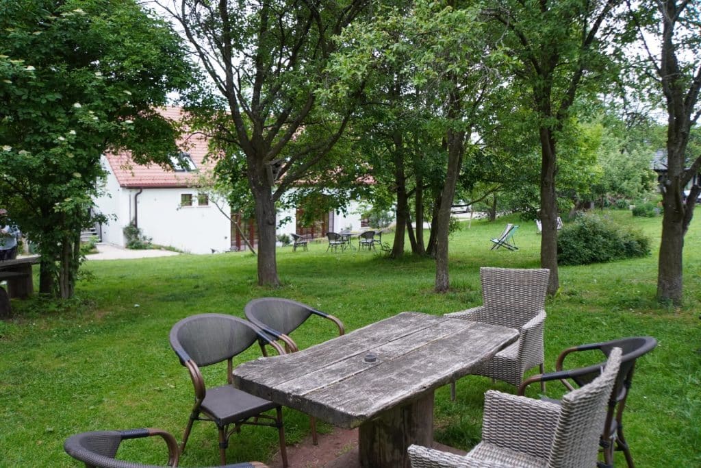 Ein rustikaler Holztisch mit verschiedenen Gartenstühlen steht im grünen Garten der Villa Greta in Niederschlesien, umgeben von schattigen Bäumen und mit Blick auf das weiße Gästehaus im Hintergrund.