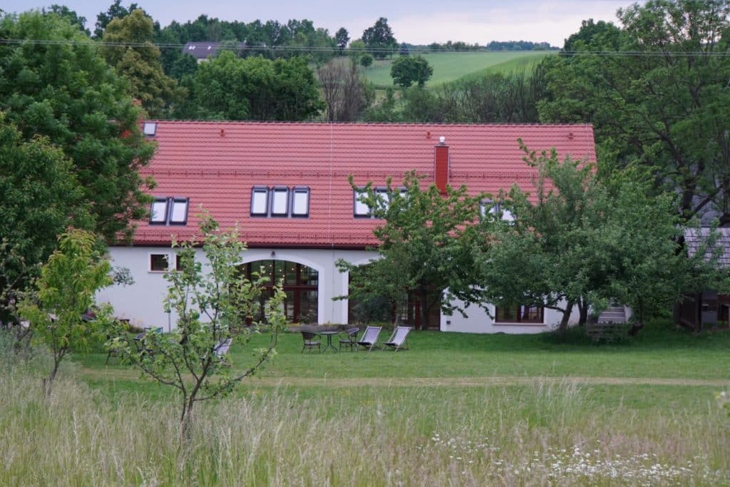Die Remise der Villa Greta in Niederschlesien präsentiert sich als weißes Gebäude mit rotem Ziegeldach, umgeben von einer weitläufigen Wiese mit Obstbäumen und Liegestühlen.