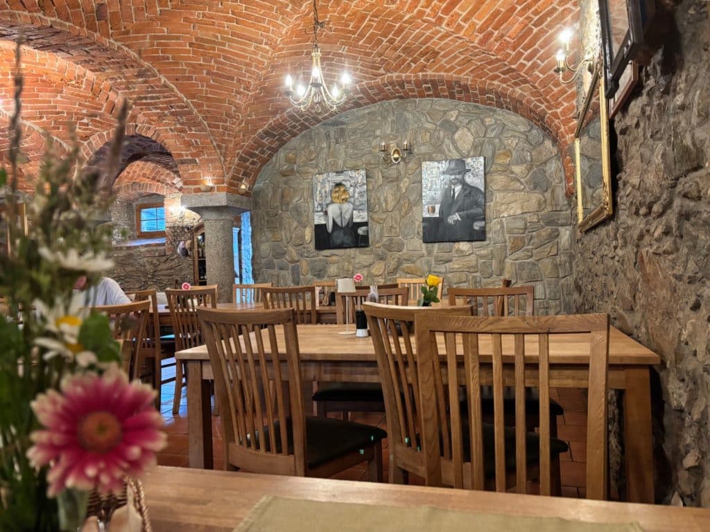 Der Innenraum des Restaurants Villa Greta in Niederschlesien zeigt einen rustikalen Gastraum mit einem markanten Ziegelgewölbe, Natursteinwänden sowie Holztischen und -stühlen. Im Vordergrund stehen unscharf fokussierte Blumen, während im Hintergrund zwei Schwarz-Weiß-Gemälde an der Steinwand hängen.