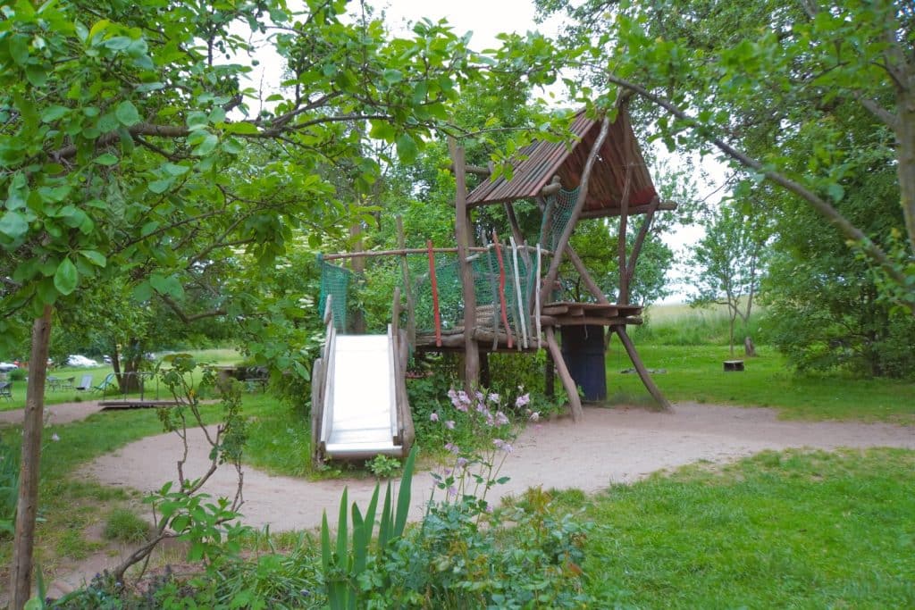 Ein rustikaler Holzspielplatz bei der Villa Greta in Niederschlesien mit einem Baumhaus auf Stelzen, einer Rutsche und einem Kletternetz inmitten einer grünen Gartenanlage.