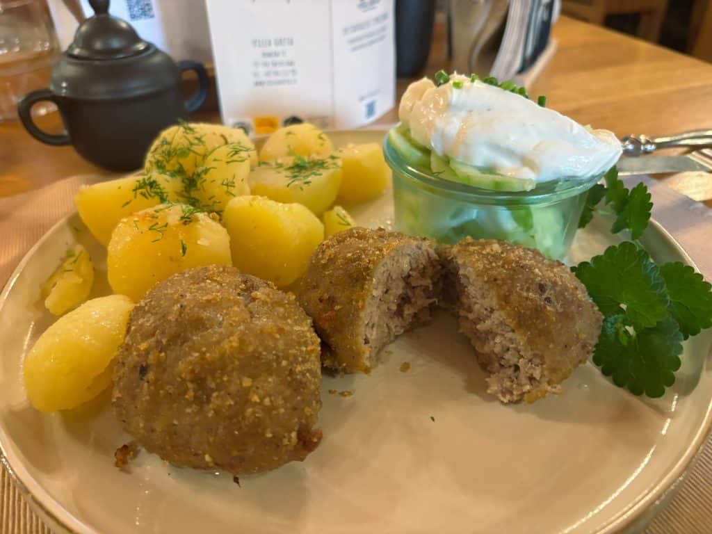 Ein Teller mit einem Tagesgericht der Villa Greta in Niederschlesien, bestehend aus zwei panierten Fleischklößchen, wovon eines halbiert ist, serviert mit Salzkartoffeln mit Dill und einem Glas Gurkensalat mit Schmand.