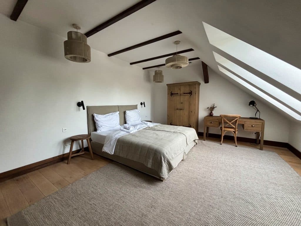 Ein helles, rustikales Dachgeschosszimmer in der Villa Greta in Niederschlesien mit einem Doppelbett, Holzmöbeln und schrägen Dachfenstern über einem Schreibtisch. Ein großer, heller Teppich liegt auf dem Holzboden unter den dunklen Deckenbalken und dekorativen Hängelampen.