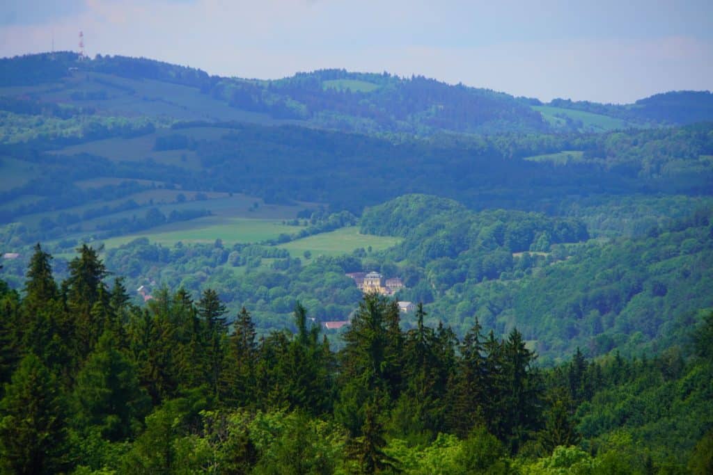 Weite Aussicht von einem Turm über die bewaldeten Hügel und grünen Täler von Niederschlesien während einer Wanderung, mit einem kleinen Dorf im Tal und einem Sendemast auf einem fernen Bergrücken.