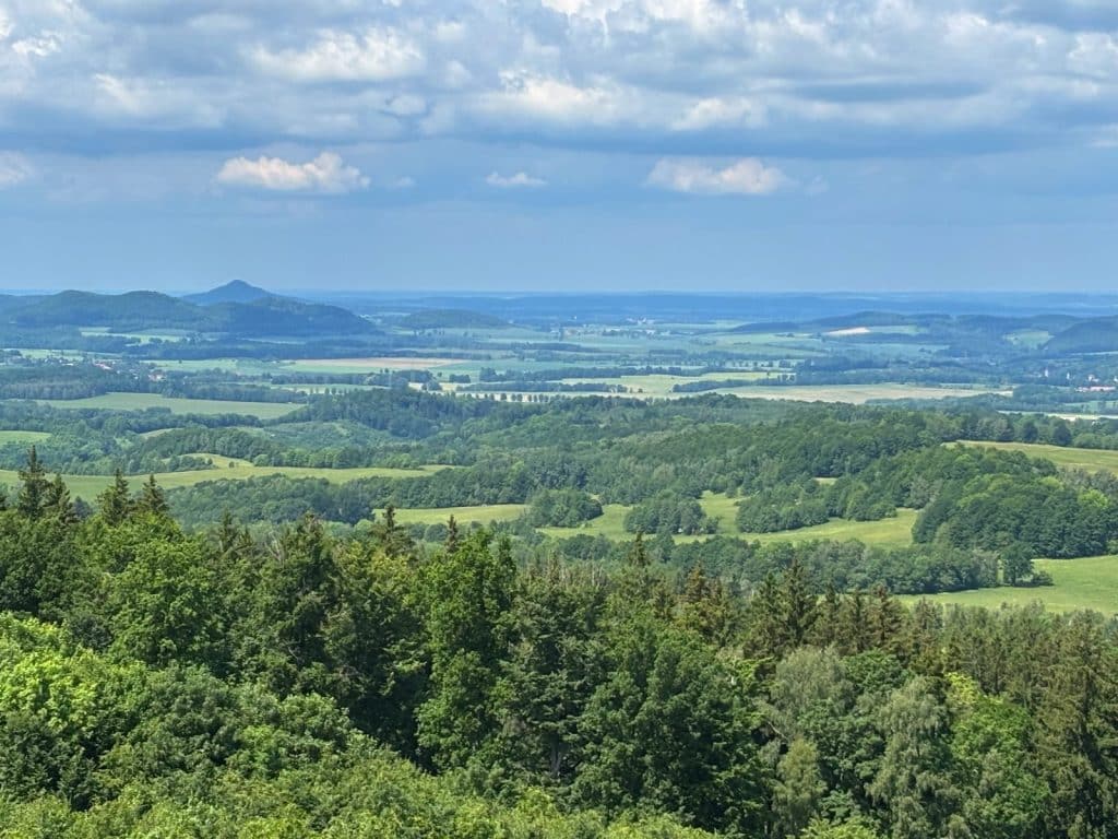 Weite Aussicht über die hügelige, bewaldete Landschaft Niederschlesiens mit grünen Wiesen und vereinzelten Bergen unter einem leicht bewölkten Sommerhimmel.