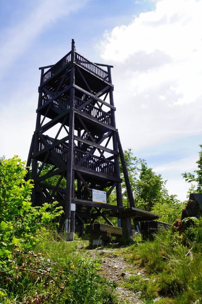 Ein hoher, hölzerner Aussichtsturm mit mehreren Plattformen steht auf einem bewaldeten Gipfel in Niederschlesien unter einem leicht bewölkten Himmel. Ein schmaler Pfad führt an einer rustikalen Sitzgruppe vorbei direkt zum Fuß der dunklen Holzkonstruktion.