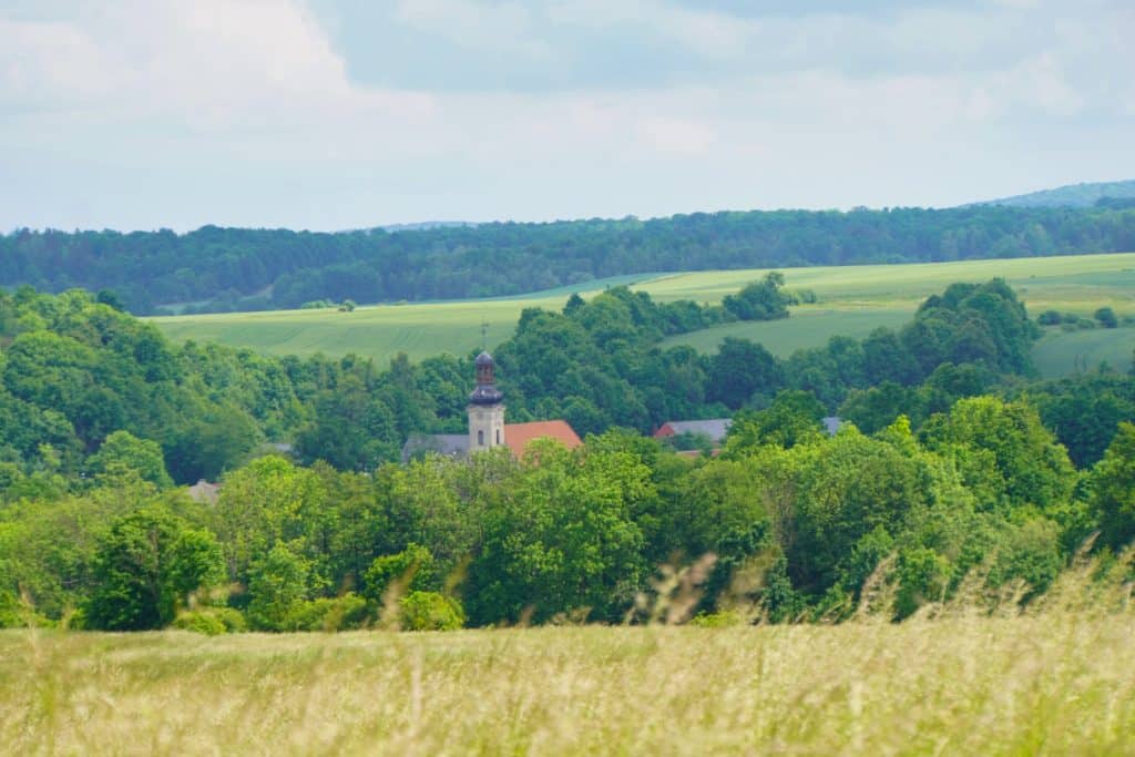 Weite, hügelige Landschaft in Niederschlesien mit Blick auf das Dorf Dobków, dessen Kirchturm und rote Dächer aus dichten grünen Wäldern und Feldern hervorragen.