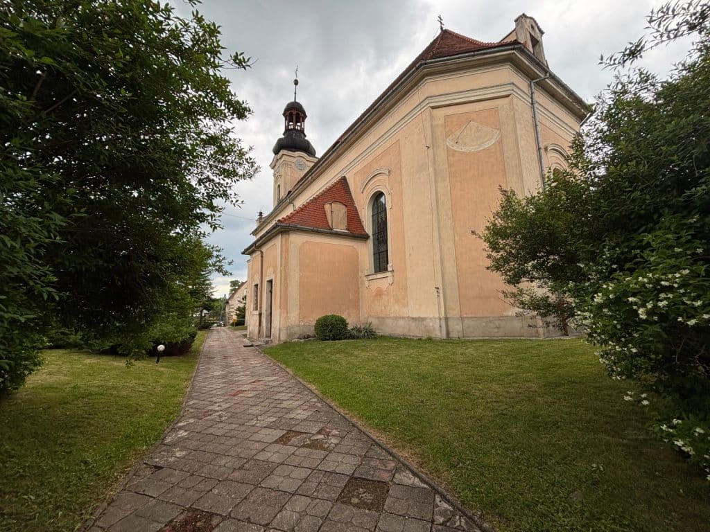 Die barocke St.-Giles-Kirche in Dobków, Niederschlesien, mit ihrer pfirsichfarbenen Fassade, einem markanten Zwiebelturm und einer Sonnenuhr an der Außenwand, gesäumt von einem gepflasterten Weg und grünen Bäumen.