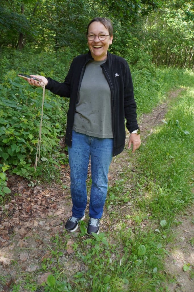 Eine lächelnde Frau mit Brille steht während einer Wanderung in Niederschlesien auf einem Waldweg, wobei ihre Kleidung durch den Regen deutlich nass geworden ist. Sie trägt eine dunkle Jacke, ein graues T-Shirt sowie feuchte Jeans und hält ein Smartphone an einer Kordel in der Hand.