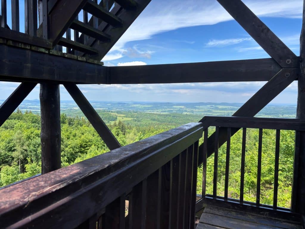 Blick von einer hölzernen Aussichtsplattform während einer Wanderung in Niederschlesien auf eine weite, grüne Wald- und Hügellandschaft unter blauem Himmel.