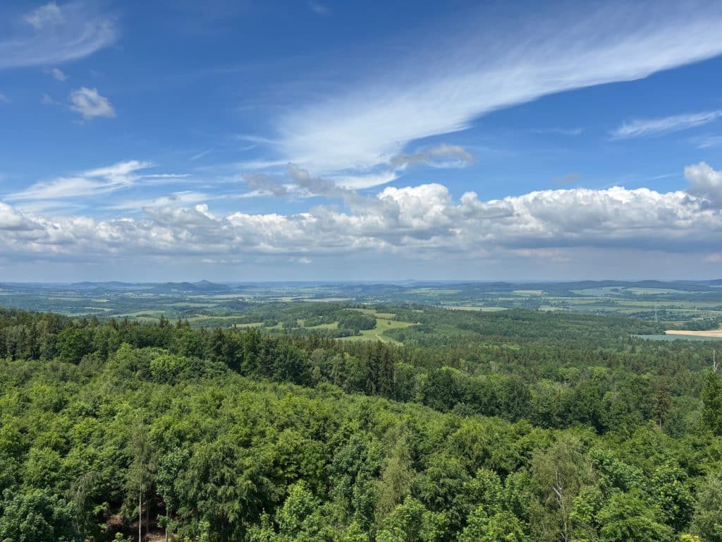 Weite Panorama-Aussicht von einem Aussichtsturm über die hügelige, bewaldete Landschaft von Niederschlesien unter einem blau-weißen Wolkenhimmel.