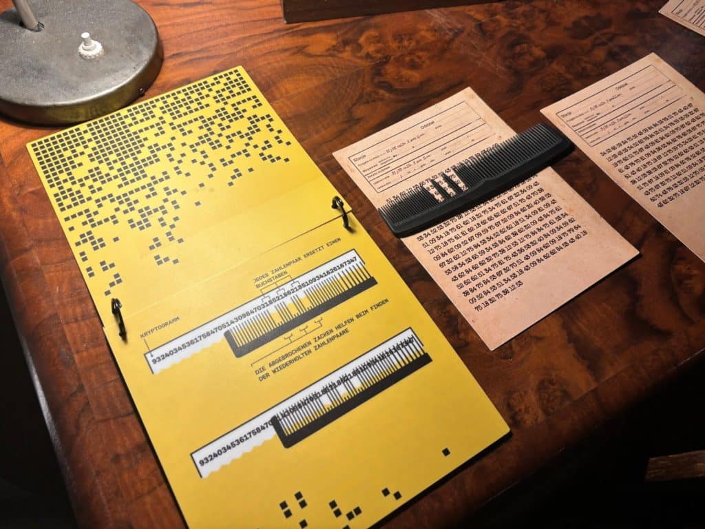 Eine Nahaufnahme zeigt ein gelbes Dokument zur Kammverschlüsselung mit Anleitungen und Diagrammen, daneben liegen ein schwarzer Kamm und zwei Papierbögen mit langen Zahlenreihen auf einer Holzoberfläche.