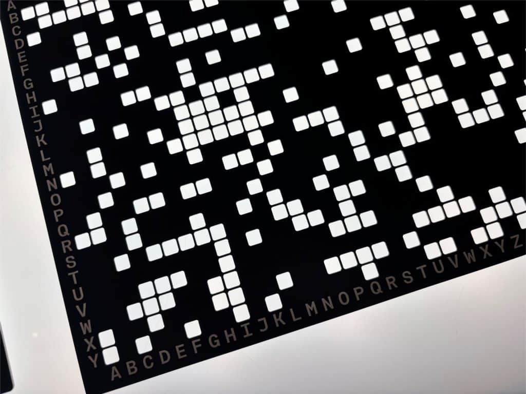 Eine Nahaufnahme der Lochplatte zeigt eine schwarze Fläche mit einem Raster aus quadratischen Löchern und alphabetischen Beschriftungen an den Rändern. Die weißen Quadrate bilden ein komplexes, unregelmäßiges Muster, das für die kryptografische Entschlüsselung verwendet wurde.