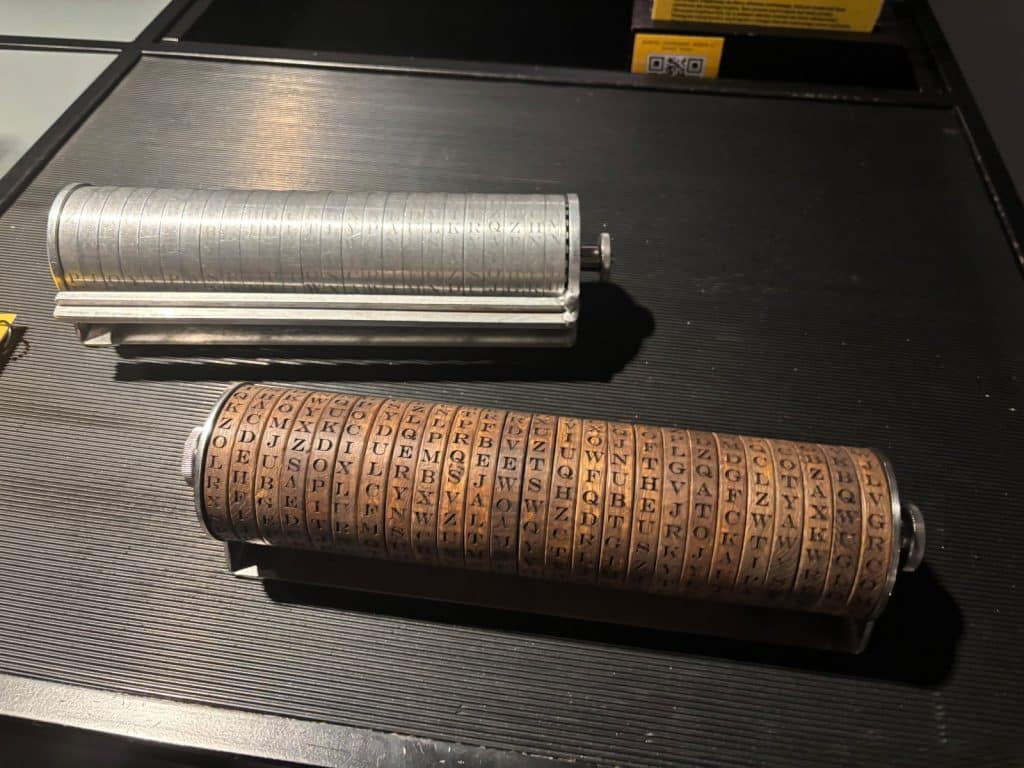 Zwei historische Buchstabenwalzen aus Metall und Holz liegen nebeneinander auf einer dunklen, geriffelten Oberfläche und zeigen verschiedene eingravierte Buchstabenfolgen auf ihren drehbaren Ringen.