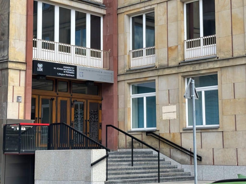 Der Eingangsbereich des Enigma-Zentrums im Collegium Martineum in Posen zeigt eine Steintreppe, die zu einer hölzernen Doppeltür unter einem schwarzen Hinweisschild der Adam-Mickiewicz-Universität führt.