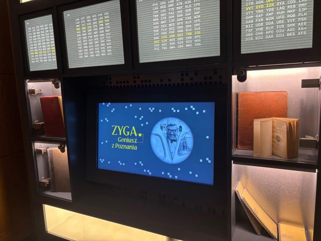 Eine Museumsausstellung im Enigma-Zentrum in Posen zeigt einen Bildschirm mit der Aufschrift „ZYGA. Geniusz z Poznania“ sowie Porträtfotos und Informationstafeln mit verschlüsselten Texten, umrahmt von historischen Büchern in beleuchteten Vitrinen.