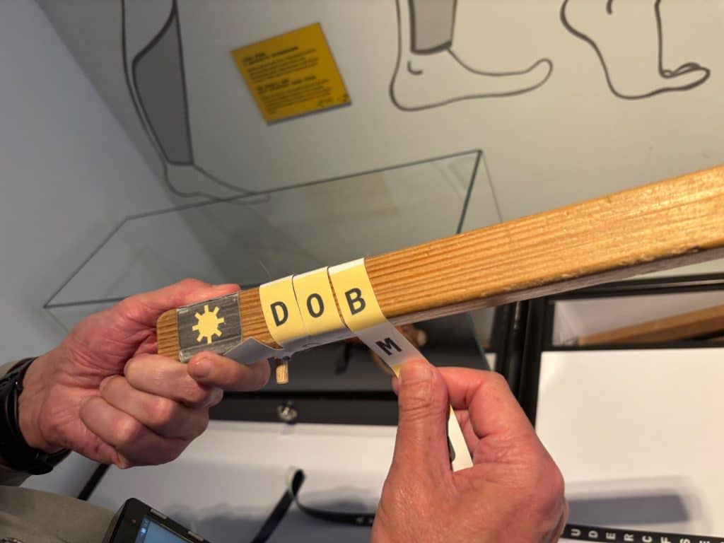 Zwei Hände demonstrieren die Holzstabmethode, bei der bedruckte Papierstreifen mit Buchstaben um einen quadratischen Holzstab gewickelt werden.