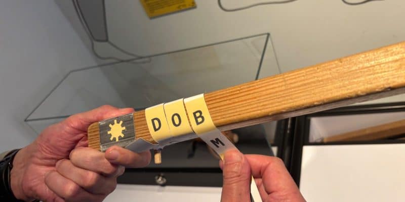 Zwei Hände demonstrieren die Holzstabmethode, bei der bedruckte Papierstreifen mit Buchstaben um einen quadratischen Holzstab gewickelt werden.