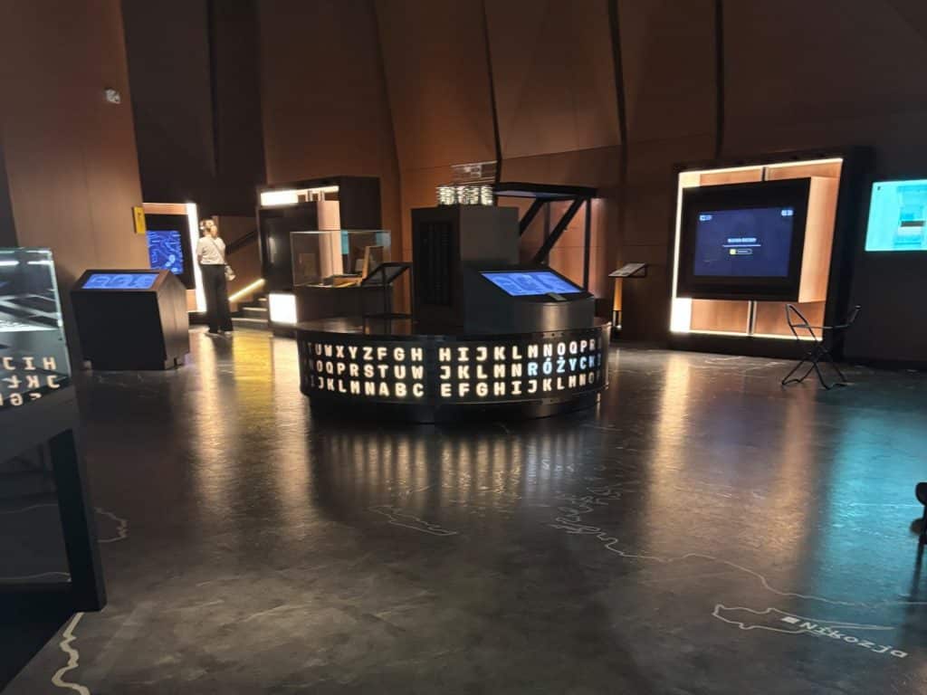 Ein modern gestalteter Ausstellungsraum im Enigma-Zentrum in Posen zeigt interaktive Stationen und eine zentrale, kreisförmige Installation mit leuchtenden Buchstabenfolgen auf einem dunklen, spiegelnden Boden. Inmitten der gedimmten Beleuchtung sind Bildschirme, Schaukästen und eine Person im Hintergrund zu sehen, während auf dem Boden geografische Umrisse und Städtenamen wie Nikosia markiert sind.
