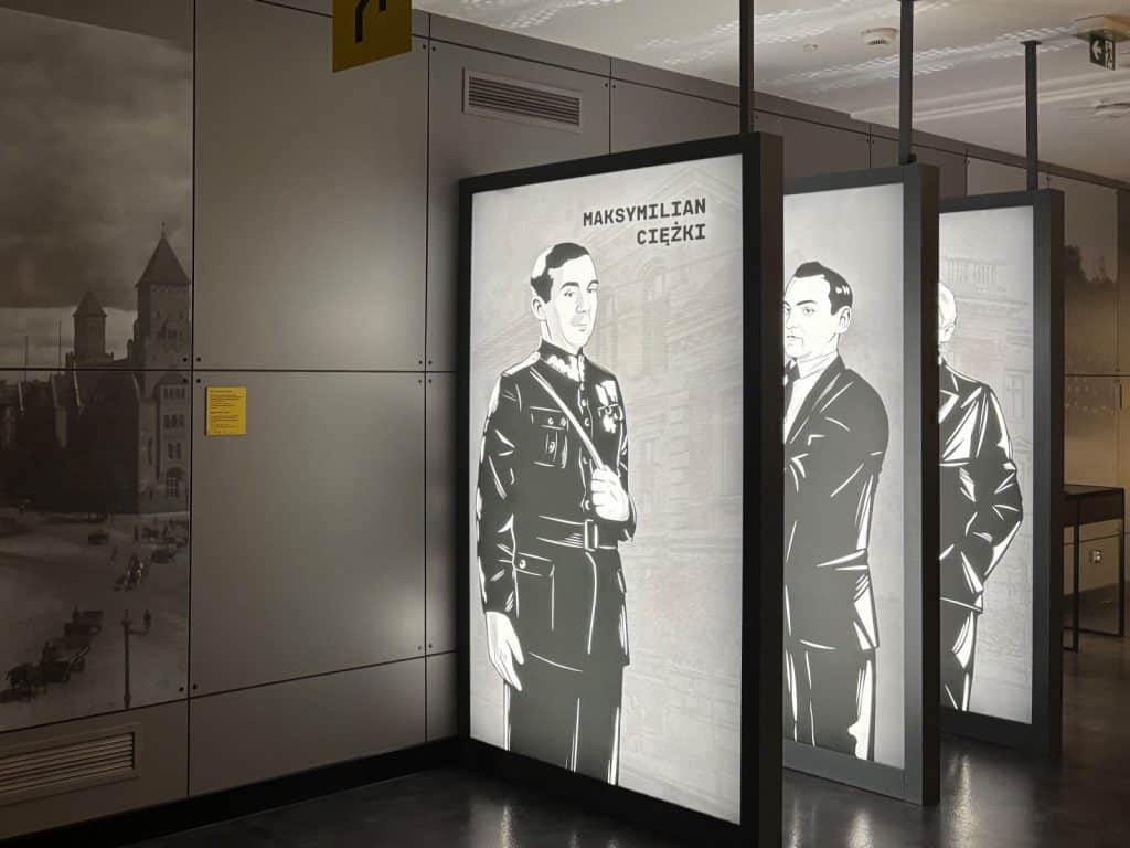 Drei beleuchtete Informationstafeln im Enigma-Museum in Posen zeigen stilisierte Porträts polnischer Codeknacker, darunter Maksymilian Ciężki in Uniform, vor einer Wand mit historischen Stadtansichten.