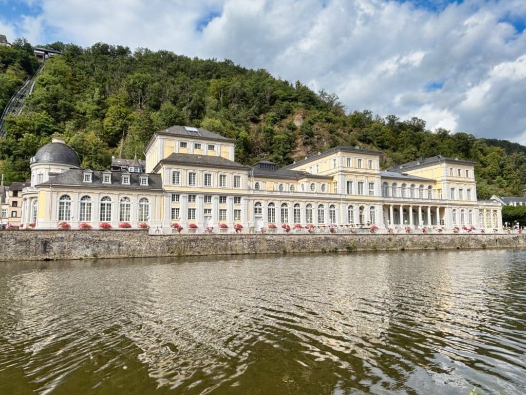 Das prachtvolle, gelb-weiße Kurhaus von Bad Ems erstreckt sich am Ufer der Lahn vor einem bewaldeten Hang, an dem eine Standseilbahn nach oben führt.