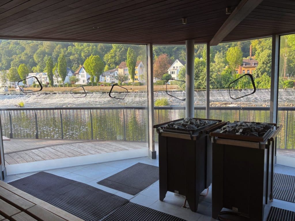 Blick aus der modernen Flusssauna der Emser Therme durch große Panoramafenster auf die Lahn, ein Wehr und die gegenüberliegenden Häuser am bewaldeten Ufer.