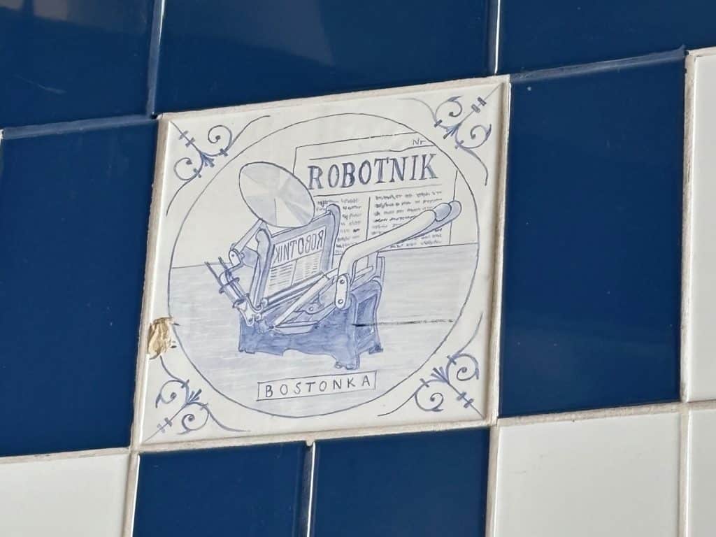 Eine quadratische, weiß-blaue Keramikfliese zeigt eine Illustration einer historischen „Bostonka“-Tiegelpresse, die eine Ausgabe der polnischen Zeitung „Robotnik“ aus Łódź druckt.
