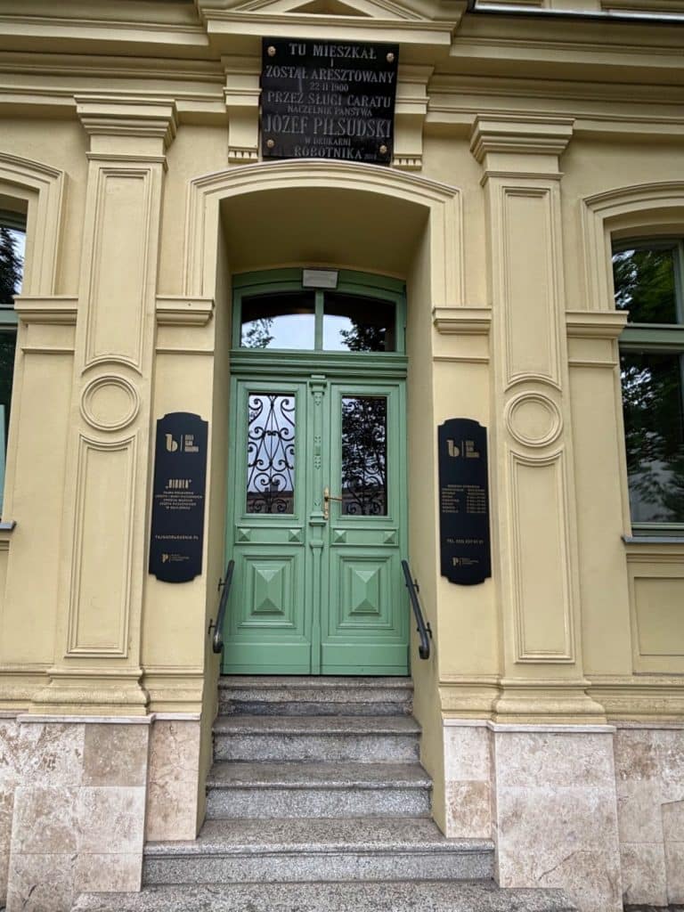 Der Eingangsbereich des Museum Bibuła Łódź zeigt eine grüne Doppeltür mit schmiedeeisernen Details, flankiert von zwei schwarzen Informationstafeln und einer Gedenktafel für Józef Piłsudski über dem Türsturz.