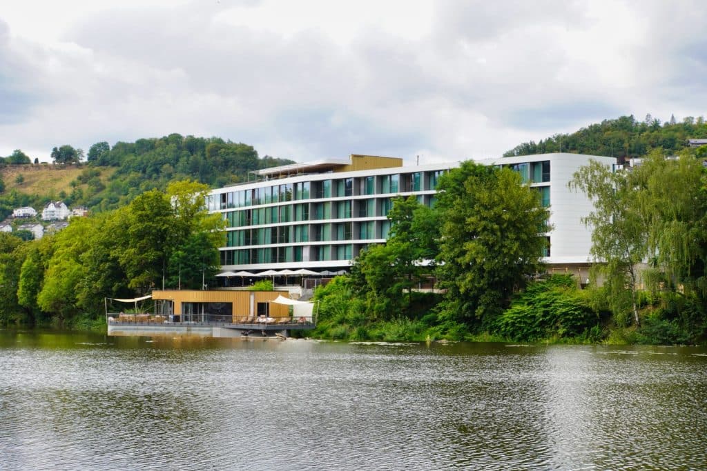 Ein modernes, mehrstöckiges Hotelgebäude mit Glasfronten in Bad Ems, das direkt am bewaldeten Flussufer liegt und über eine vorgelagerte Terrasse am Wasser verfügt.