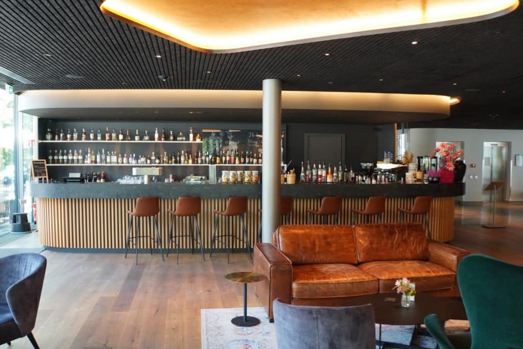 Die moderne Hotelbar in Bad Ems besticht durch einen langen Tresen mit Holzlamellen, braune Barhocker und einen gemütlichen Loungebereich mit einem braunen Ledersofa im Vordergrund.