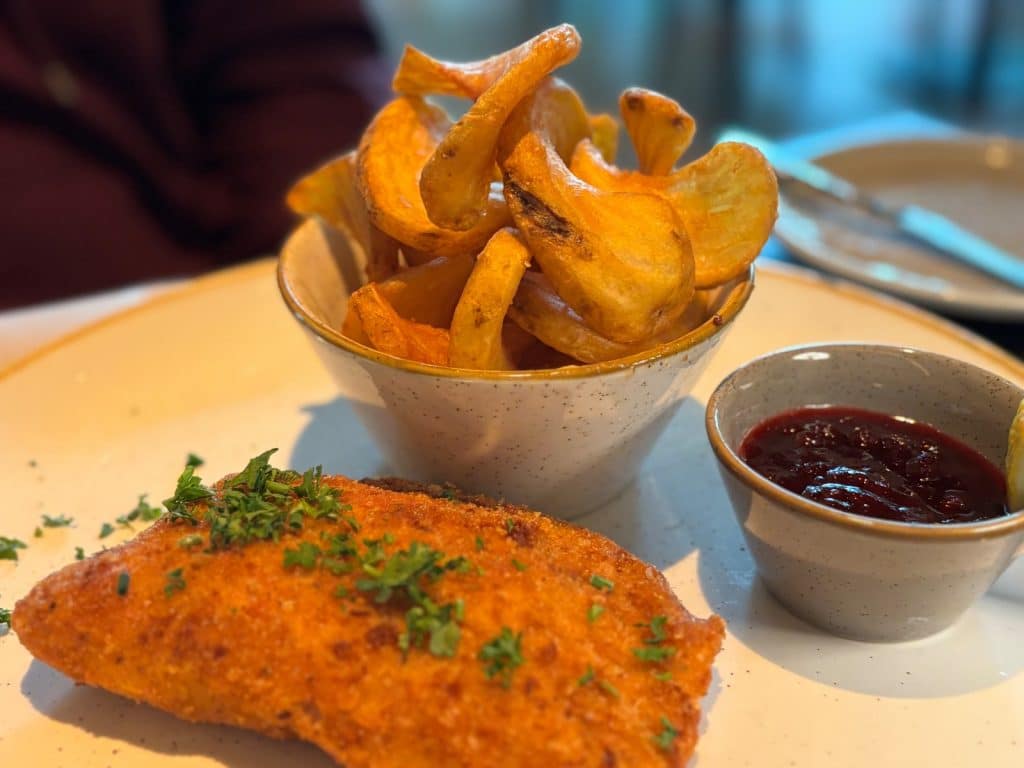 Ein goldbraun paniertes Cordon bleu, garniert mit frischer Petersilie, wird zusammen mit geschwungenen Kartoffelspalten in einer Schüssel und einer dunklen Preiselbeersauce in einem Hotel in Bad Ems serviert.