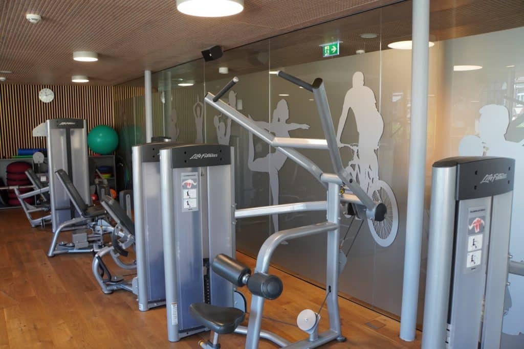 Der moderne Fitnessraum im Hotel in Bad Ems ist mit verschiedenen Kraftgeräten von Life Fitness, Holzboden und einer Glaswand mit sportlichen Silhouetten ausgestattet.