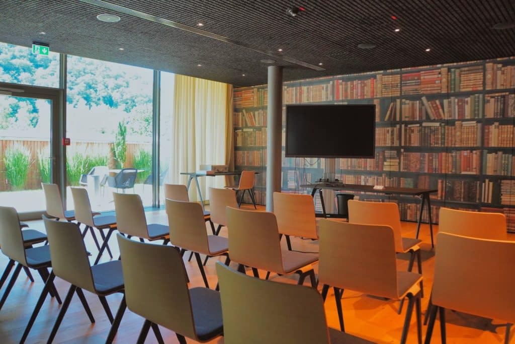 Ein modern gestalteter Konferenzraum im Bad Ems Hotel mit mehreren Reihen schlichter Holzstühle, die auf einen großen Bildschirm vor einer markanten Tapete in Bücherregal-Optik ausgerichtet sind. Eine große Glasfront gibt den Blick auf eine Terrasse und grüne Hügel frei.