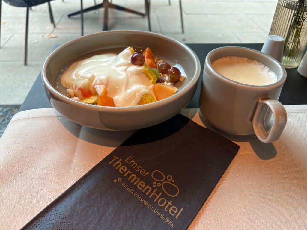Ein Frühstück im Emser ThermenHotel in Bad Ems mit einer Schale Müsli mit Joghurt und frischen Früchten neben einer Tasse Kaffee auf einer Serviette mit Hotel-Logo.
