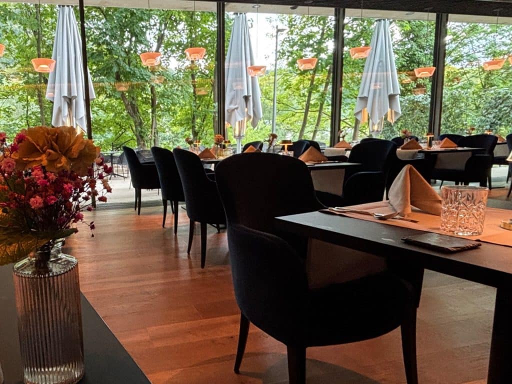 Ein elegant eingerichtetes Restaurant im Bad Ems Hotel mit dunklen Polstersesseln an eingedeckten Tischen vor einer großen Fensterfront mit Blick ins Grüne. Im Vordergrund steht eine Vase mit bunten Blumen auf einem Tisch, während draußen geschlossene weiße Sonnenschirme zu sehen sind.