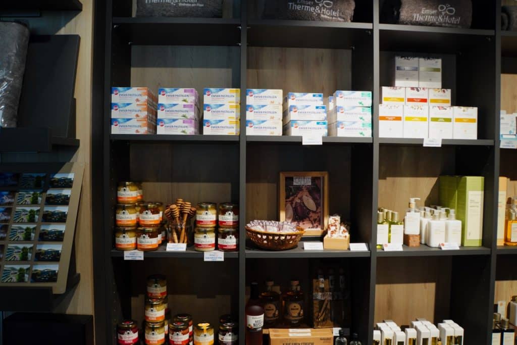Ein gut sortiertes Verkaufsregal im Shop des Emser Therme & Hotels präsentiert verschiedene regionale Produkte wie Honiggläser, Pastillen, Pflegeartikel und Souvenirs. Die Waren sind ordentlich in dunklen Holzfächern angeordnet, ergänzt durch Postkarten auf der linken Seite.