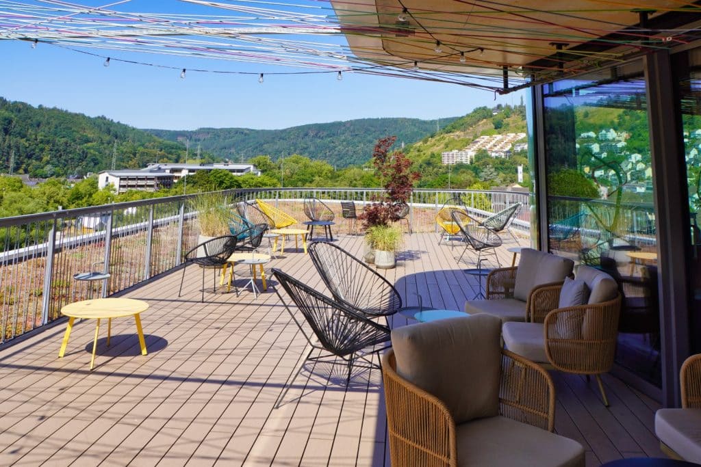 Die sonnige Skylounge-Terrasse eines Hotels in Bad Ems bietet moderne Loungemöbel, farbenfrohe Acapulco-Stühle und einen weiten Panoramablick über die bewaldeten Hügel und Gebäude der Stadt.