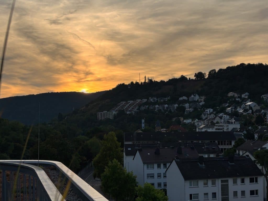 Blick von einem Balkon auf den Sonnenuntergang über den bewaldeten Hängen und Wohngebäuden von Bad Ems, während der Himmel in warmen Orange- und Gelbtönen leuchtet.
