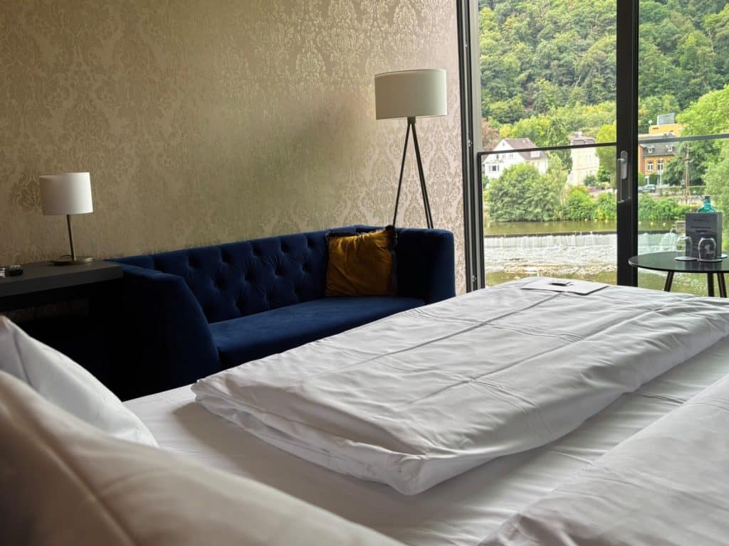 Ein gemütliches Hotelzimmer in Bad Ems mit einem weißen Bett im Vordergrund, einem blauen Samtsofa an einer gemusterten Wand und einem großen Fenster, das den Blick auf die Lahn und bewaldete Hügel freigibt.