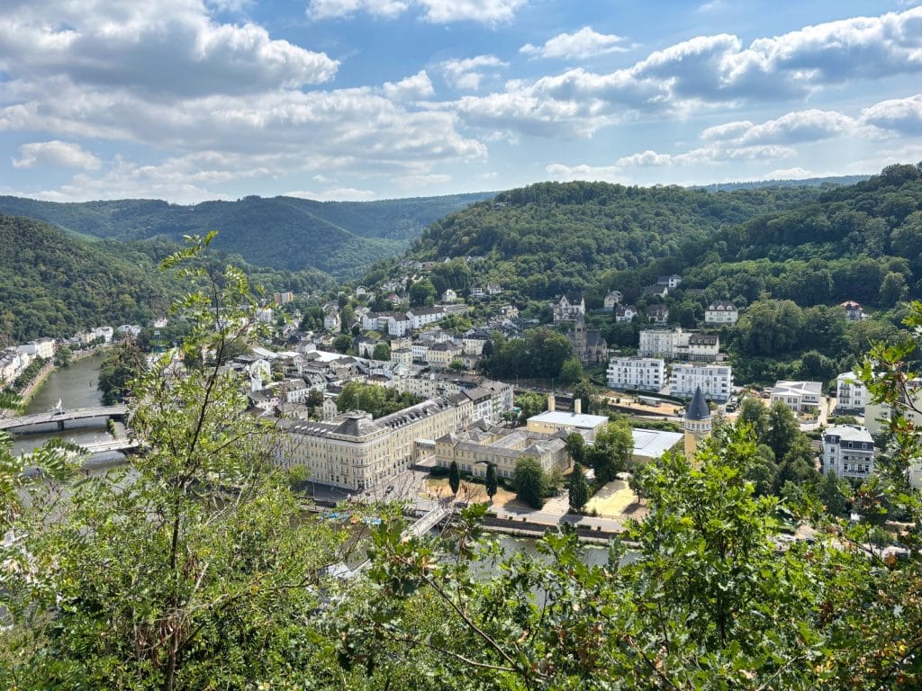 Weitwinkel-Aussicht vom Vogler Aussichtspunkt auf das im Flusstal der Lahn gelegene Bad Ems mit seinen historischen Kurgebäuden, umgeben von dicht bewaldeten Hügeln unter einem leicht bewölkten Sommerhimmel.