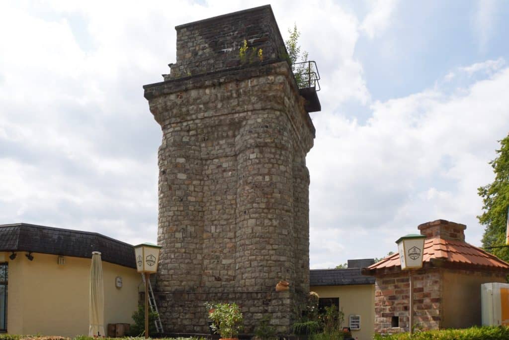 Ein massiver, steinerner Bismarckturm in Bad Ems ragt hinter einem flachen Gebäude in den bewölkten Himmel, wobei auf seiner oberen Plattform kleine Pflanzen wachsen.