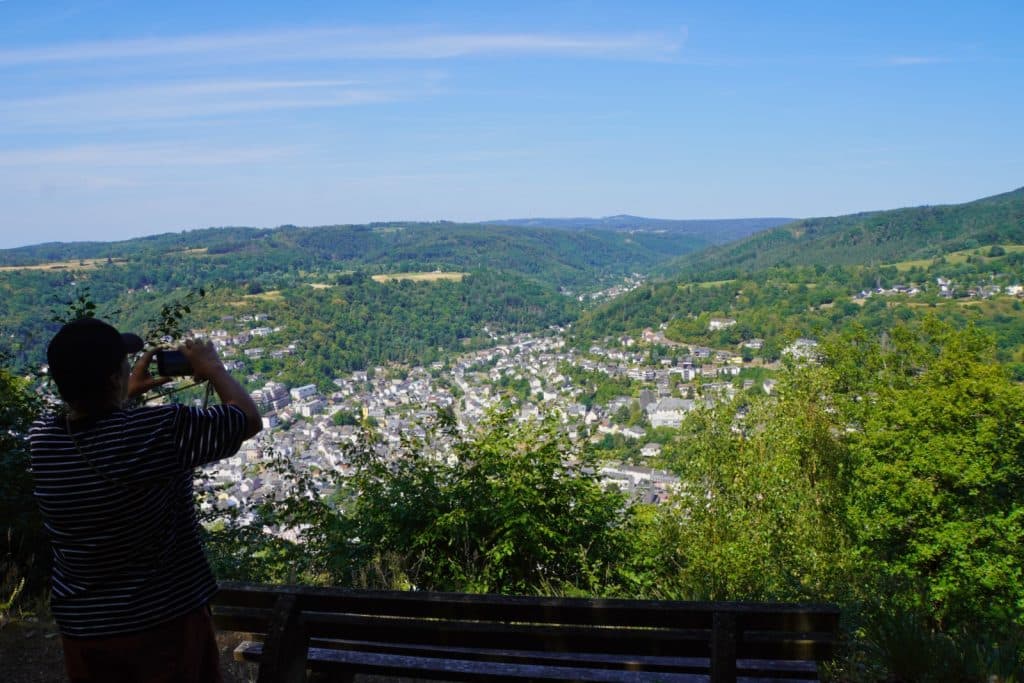 Ein Wanderer steht auf dem Malberg bei Bad Ems neben einer Holzbank und fotografiert den weiten Ausblick über die Stadt im Tal und die bewaldeten Hügel.