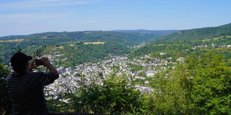 Ein Wanderer steht auf dem Malberg bei Bad Ems neben einer Holzbank und fotografiert den weiten Ausblick über die Stadt im Tal und die bewaldeten Hügel.
