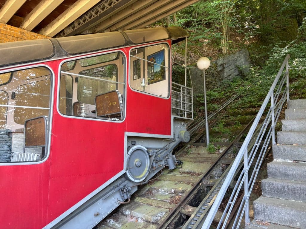 Ein roter Wagen der historischen Malbergbahn in Bad Ems steht auf den steilen Schienen der bewachsenen Strecke neben einer Steintreppe.