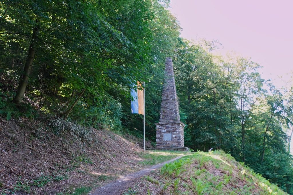 Ein Waldweg führt zur Henriettensäule bei Bad Ems, einem steinernen Obelisken auf dem Malberg, neben dem eine gelb-blaue Flagge weht.
