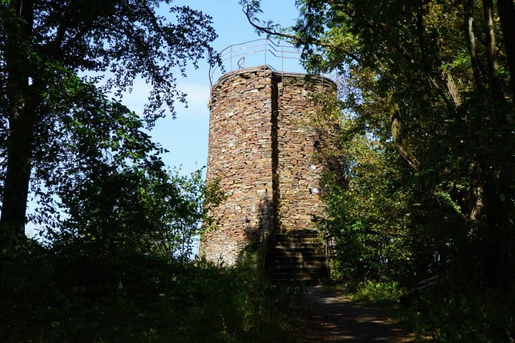 Ein runder, aus Natursteinen gemauerter Aussichtsturm auf dem Malberg bei Bad Ems, der von dichten grünen Bäumen eingerahmt wird und über eine Steintreppe zugänglich ist.