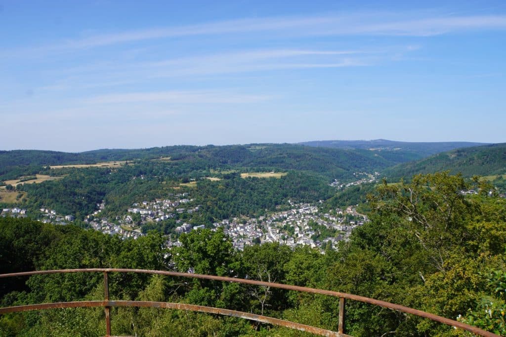 Blick vom Malbergturm bei Bad Ems über ein bewaldetes Tal auf die Stadt und die umliegenden grünen Hügel unter blauem Himmel. Im Vordergrund ist ein rostiges Metallgeländer einer Aussichtsplattform zu sehen.