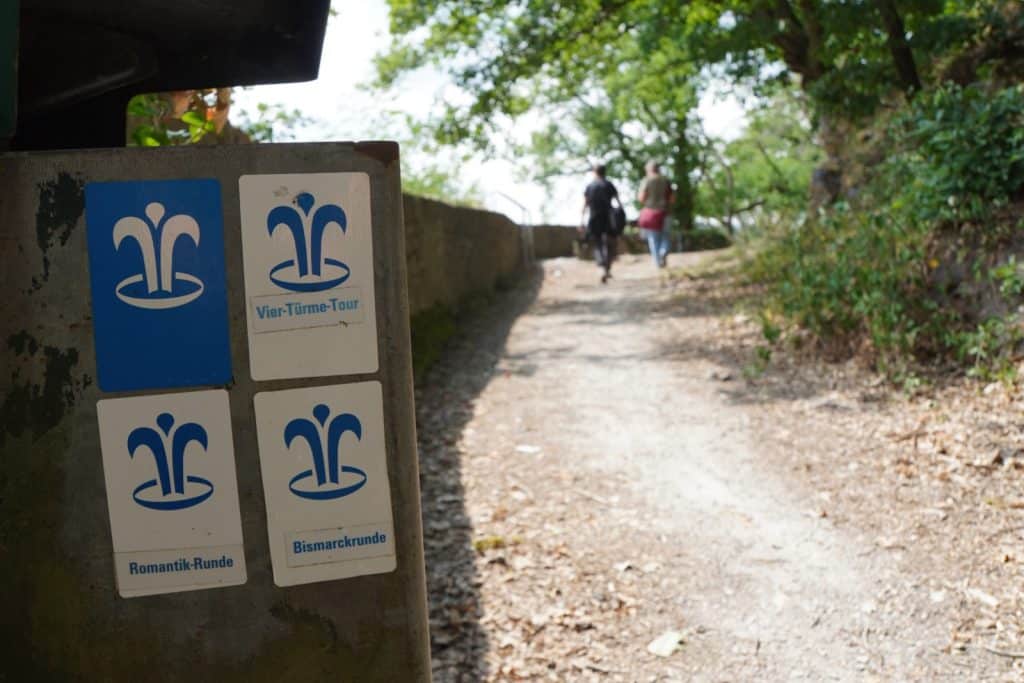 Wanderschilder am Wegesrand weisen auf verschiedene Wanderwege bei Bad Ems hin, darunter die Vier-Türme-Tour, die Romantik-Runde und die Bismarckrunde, während im Hintergrund zwei Personen auf einem Waldweg spazieren.
