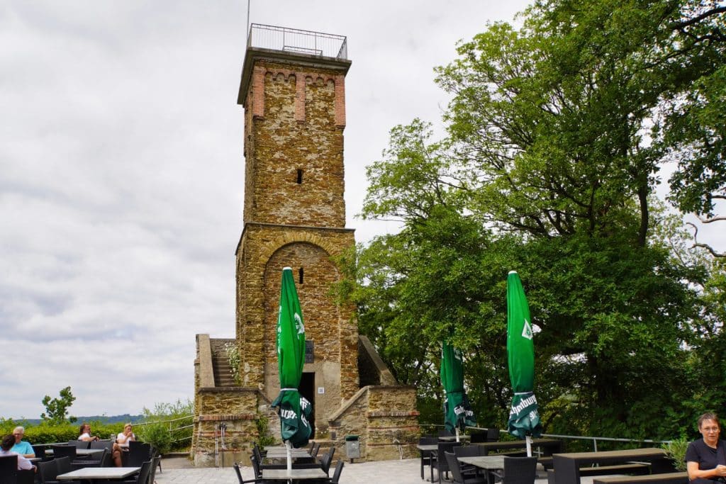Ein hoher, historischer Steinturm, der Concordiaturm bei Bad Ems, ragt neben einer Außenterrasse mit Tischen, Stühlen und grünen Sonnenschirmen unter Bäumen empor.