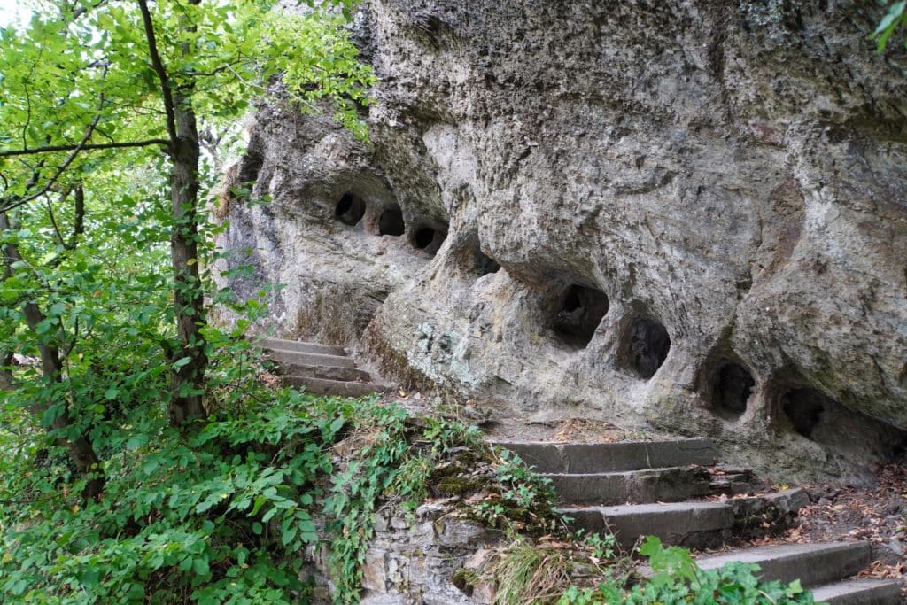 Eine Treppe aus Stein führt im Wald an den Heinzelmannhöhlen bei Bad Ems vorbei, die als kleine, runde Öffnungen in einer zerklüfteten Felswand zu sehen sind.