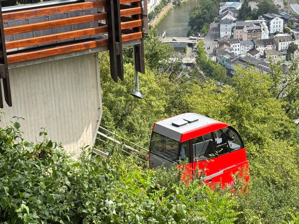 Eine rote Kabine der Kurwaldbahn in Bad Ems fährt einen bewaldeten Hang hinauf, während im Hintergrund die Stadt und die Lahn im Tal sichtbar sind.